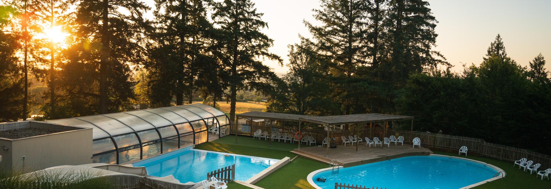 A view of the aquatic area featuring a covered heated pool, an outdoor pool, a toddler paddling pool with small slides, and surrounding sun loungers, all set against tall trees at sunset.