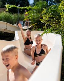 Children sliding down a water slide in a nature-filled outdoor aquatic area, smiling and having fun in the sunshine.