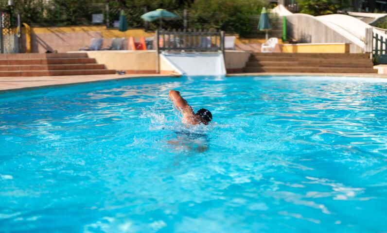 A person swimming in a large outdoor heated pool with a slide and sun terrace visible in the background on a sunny day.