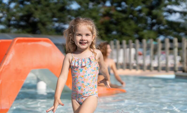Child playing in a shallow splash pool beside a small orange slide, enjoying a sunny moment in the campsite’s aquatic area.