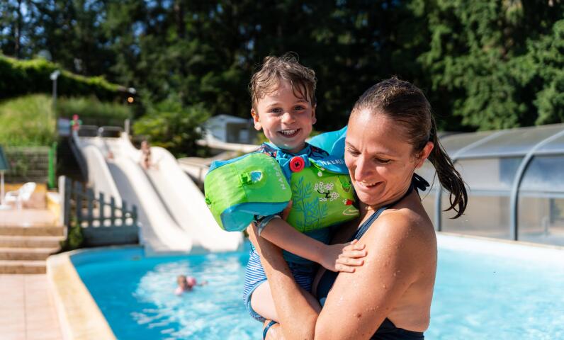 An adult holding a child wearing floaties beside a heated outdoor pool, with a water slide visible in the background on a sunny day.