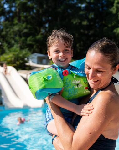 An adult holding a child wearing floaties beside a heated outdoor pool, with a water slide visible in the background on a sunny day.