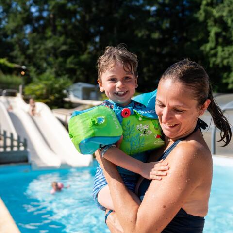 An adult holding a child wearing floaties beside a heated outdoor pool, with a water slide visible in the background on a sunny day.