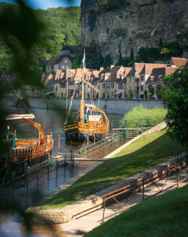 Traditional wooden gabarre boats moored along the Dordogne River in La Roque-Gageac, with stone houses and towering cliffs in the background.