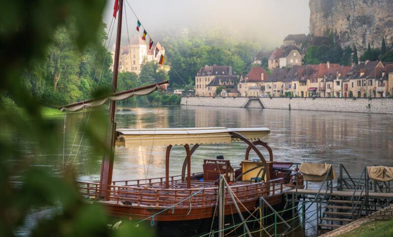 Traditional wooden gabarre moored on the Dordogne River, with the stone houses and cliffs of La Roque-Gageac in a soft morning mist.