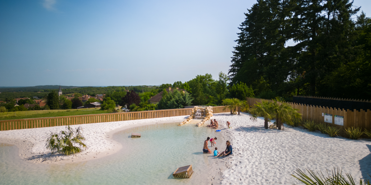 Panoramic view of a sandy lagoon with sun loungers and families relaxing in a natural aquatic area, surrounded by greenery in a calm Dordogne Valley setting.