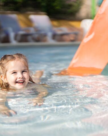 Smiling young child playing in a shallow paddling pool with a small slide, in a safe and family-friendly aquatic area.