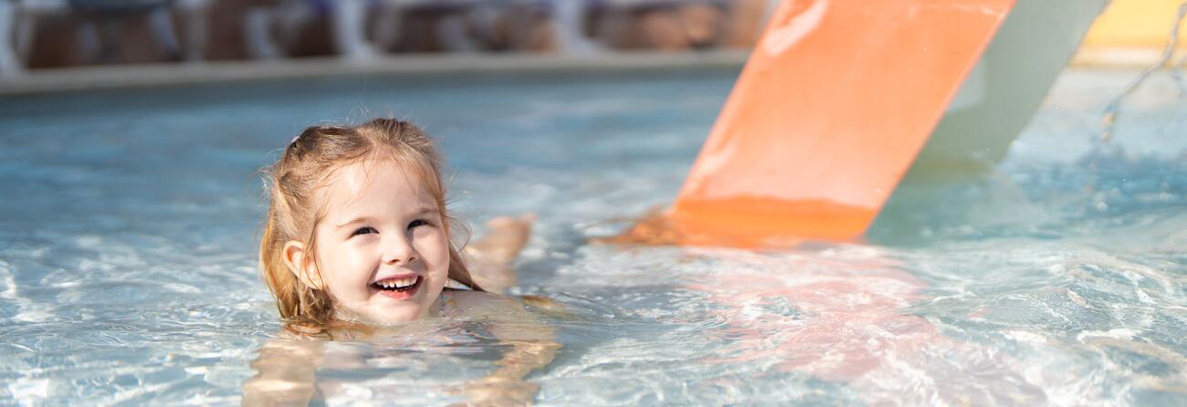 Smiling young child playing in a shallow paddling pool with a small slide, in a safe and family-friendly aquatic area.