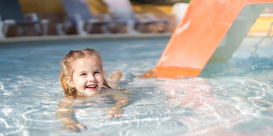 Smiling young child playing in a shallow paddling pool with a small slide, in a safe and family-friendly aquatic area.