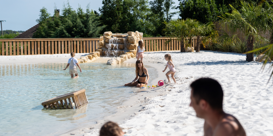 Family relaxing and playing by the natural lagoon pool with sandy beach at Camping Le Séquoïa in the Dordogne Valley.