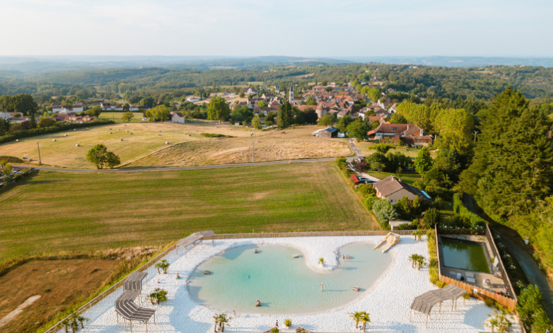 Aerial view of the natural lagoon pool surrounded by countryside at Camping Le Séquoïa in Payrac, in the heart of the Lot and Dordogne Valley.