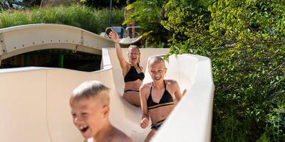 Enfants descendant un toboggan aquatique dans un parc aquatique en pleine nature, sous le soleil, créant une ambiance familiale et joyeuse.