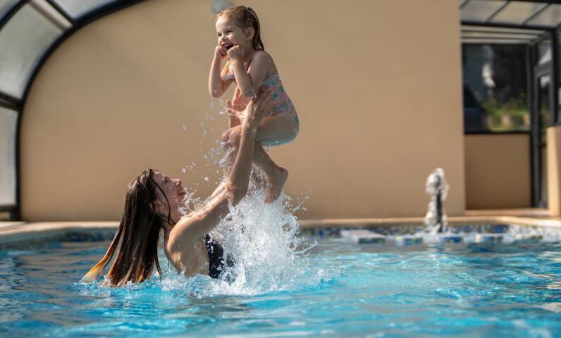 Un adulte jouant avec un enfant dans une piscine intérieure chauffée, créant des éclaboussures et un moment de joie sous une structure couverte lumineuse.