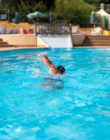 Personne nageant dans une large piscine extérieure chauffée, avec un toboggan et une terrasse en arrière-plan, sous un ciel ensoleillé.
