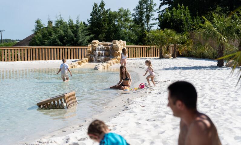 Familles et enfants jouant dans un lagon peu profond au sable blanc, avec une petite cascade rocheuse et une ambiance naturelle ensoleillée.