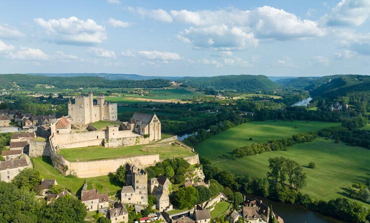 Vue aérienne du château de Beynac dominant la Dordogne, entouré de maisons en pierre et de paysages verdoyants, avec la rivière serpentant à travers la vallée.