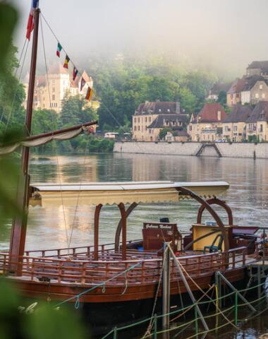 Gabare traditionnelle amarrée sur la Dordogne, avec les maisons en pierre de La Roque-Gageac et les falaises en arrière-plan dans une ambiance matinale brumeuse.