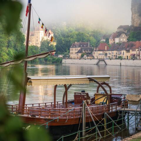 Gabare traditionnelle amarrée sur la Dordogne, avec les maisons en pierre de La Roque-Gageac et les falaises en arrière-plan dans une ambiance matinale brumeuse.
