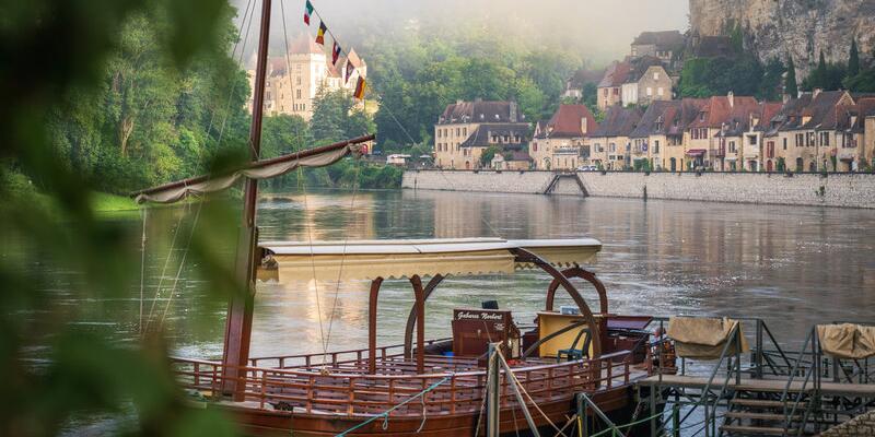 Gabare traditionnelle amarrée sur la Dordogne, avec les maisons en pierre de La Roque-Gageac et les falaises en arrière-plan dans une ambiance matinale brumeuse.