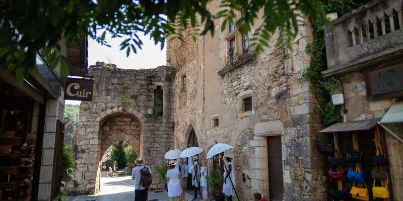 Scène de rue à Rocamadour avec des bâtiments en pierre, une ancienne porte médiévale et des visiteurs se promenant sous une lumière estivale dans ce village emblématique du Lot.