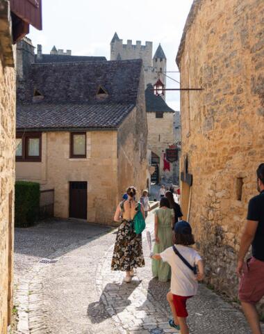 Famille marchant dans une ruelle en pierre à Beynac, entourée de maisons médiévales et baignée de lumière estivale, dans l’un des plus beaux villages de Dordogne.