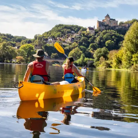Famille faisant du canoë sur la rivière Dordogne, pagayant dans une eau calme entourée de verdure, avec un château perché visible en arrière-plan.