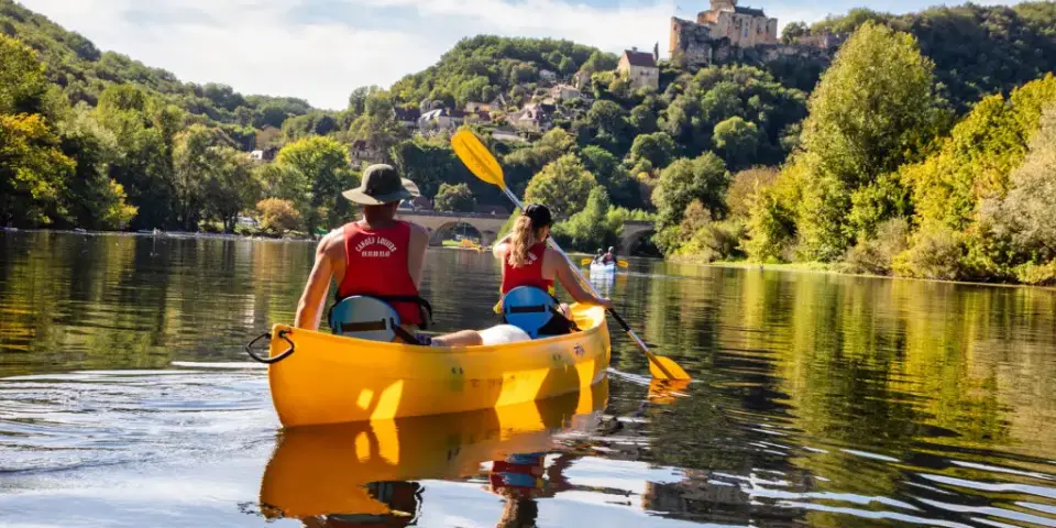 Famille faisant du canoë sur la rivière Dordogne, pagayant dans une eau calme entourée de verdure, avec un château perché visible en arrière-plan.