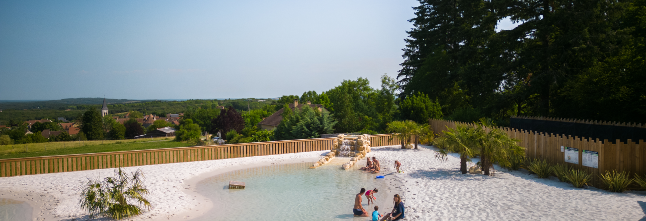 Vue panoramique d’un lagon sableux avec transats et familles profitant d’un espace aquatique naturel, entouré de verdure, dans un cadre calme et ensoleillé en Dordogne.