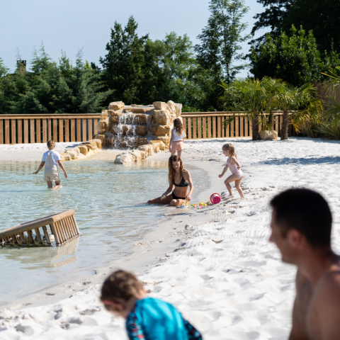 Familie ontspant en speelt bij het natuurlijke lagunezwembad met zandstrand op Camping Le Séquoïa in de Dordognevallei.