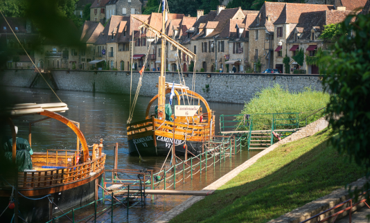 Traditionele houten gabares aangemeerd langs de rivier de Dordogne in La Roque-Gageac, met stenen huizen en hoge kliffen op de achtergrond.
