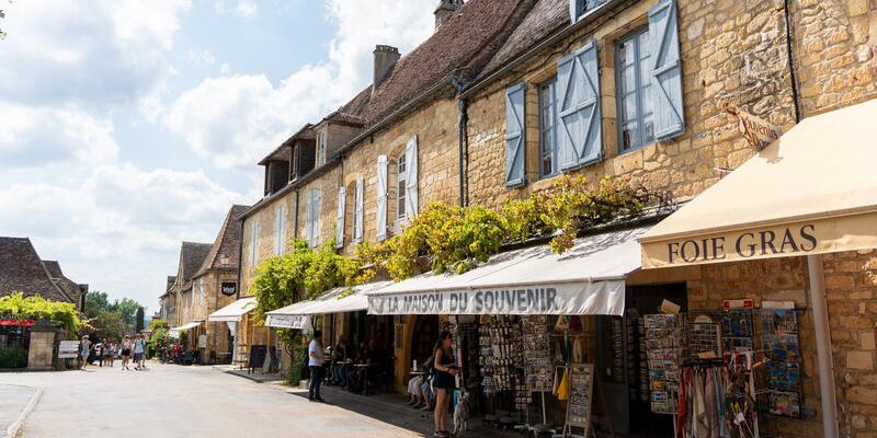 Hoofdstraat van Domme met goudkleurige stenen huizen, lokale winkels en schaduwrijke luifels onder een heldere hemel in dit bekende heuveldorp van de Dordogne.