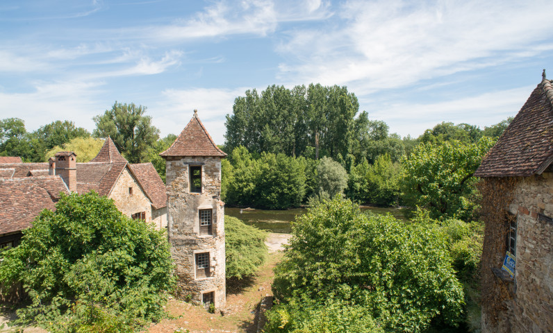 Uitzicht op een stenen gebouw met een karakteristieke toren in Carennac, omringd door groene natuur in een rustige omgeving langs de rivier.