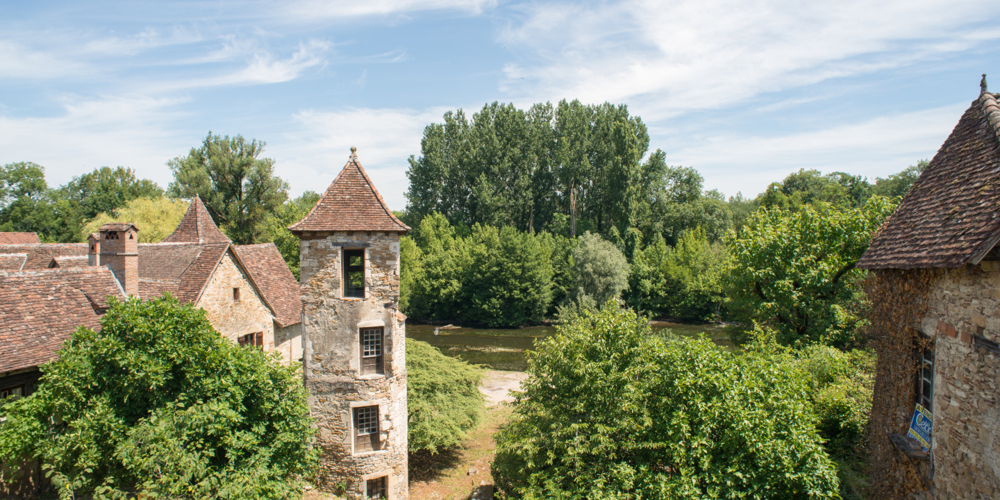 Uitzicht op een stenen gebouw met een karakteristieke toren in Carennac, omringd door groene natuur in een rustige omgeving langs de rivier.