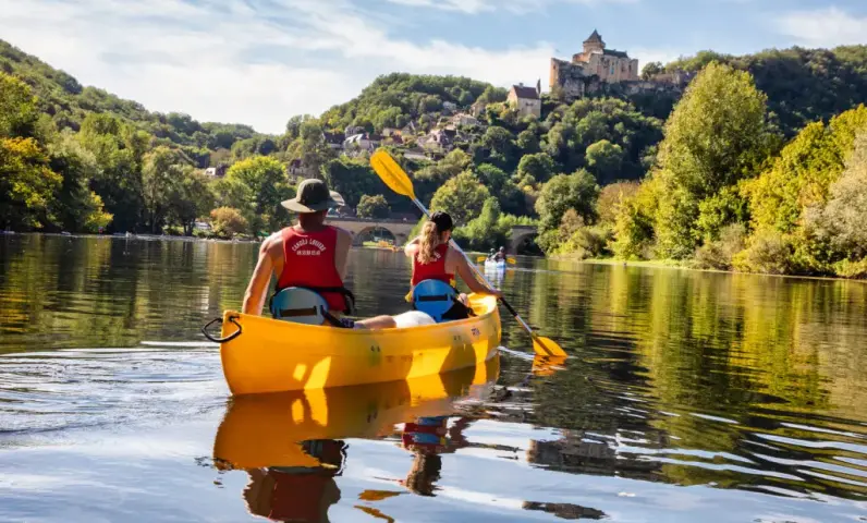 Gezin dat kano vaart op de rivier de Dordogne, peddelend door rustig water met groene oevers en een kasteel op de heuvel op de achtergrond.