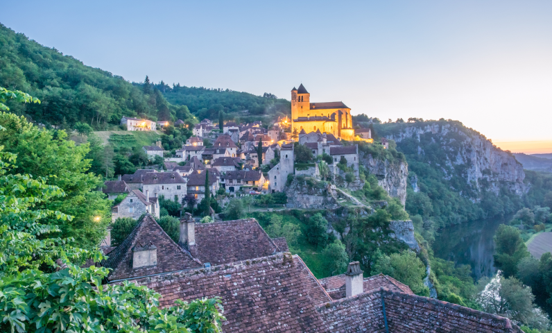 Panoramisch uitzicht op Saint-Cirq-Lapopie bij zonsondergang, met stenen huizen, de verlichte kerk en de hoge rotsen boven de Lotvallei.