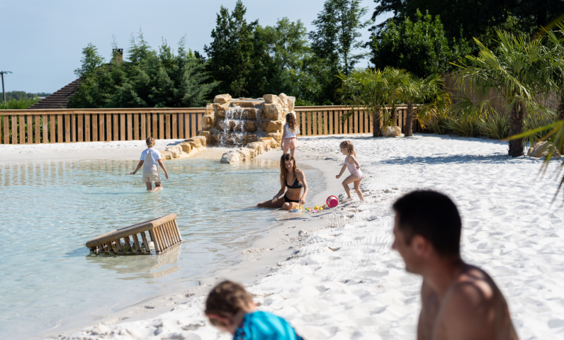 Familie ontspant en speelt bij het natuurlijke lagunezwembad met zandstrand op Camping Le Séquoïa in de Dordognevallei.